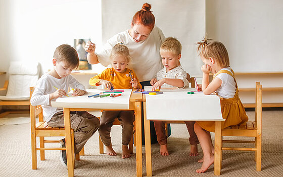 Fröhliche Lehrerin mit einer Gruppe niedlicher Vorschulkinder am Tisch, die mit bunten Filzstiften auf Papier zeichnen