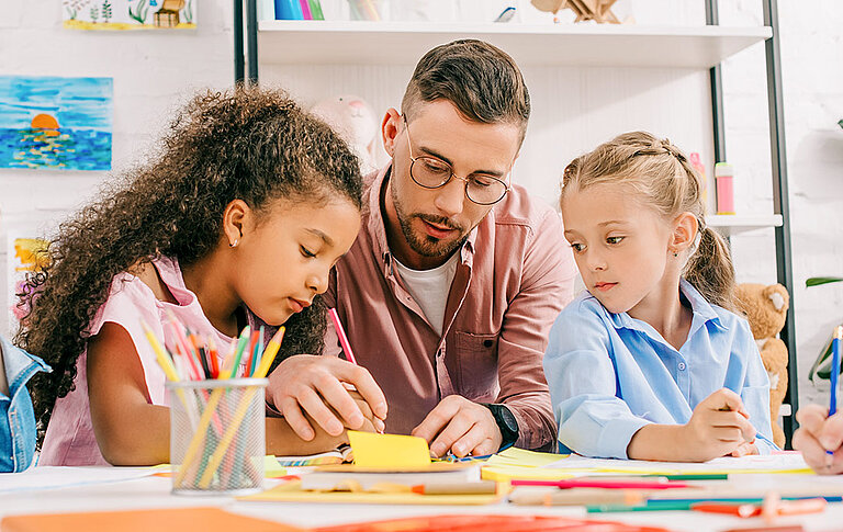 Lehrer mit Brille hilft multikulturellen Vorschulkindern beim Zeichnen am Tisch im Klassenzimmer
